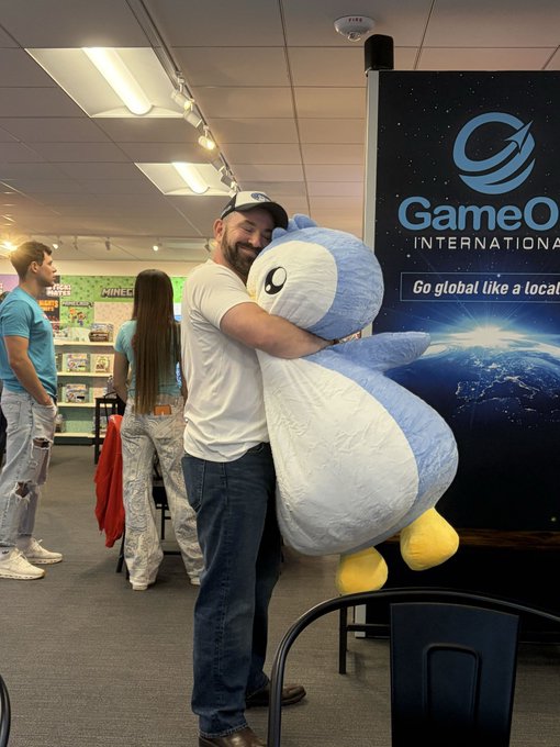 A bearded man in a white t-shirt and cap joyfully hugs a large blue and white penguin plushie at a GameOut International event, with a banner reading "Go global like a local" in the background, perfectly capturing the affection expressed for the Pudgy Penguins brand.