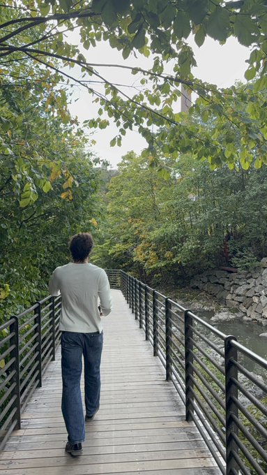 A person wearing a white shirt and jeans walking across a wooden bridge with black metal railings, surrounded by lush green trees and a stream below.
