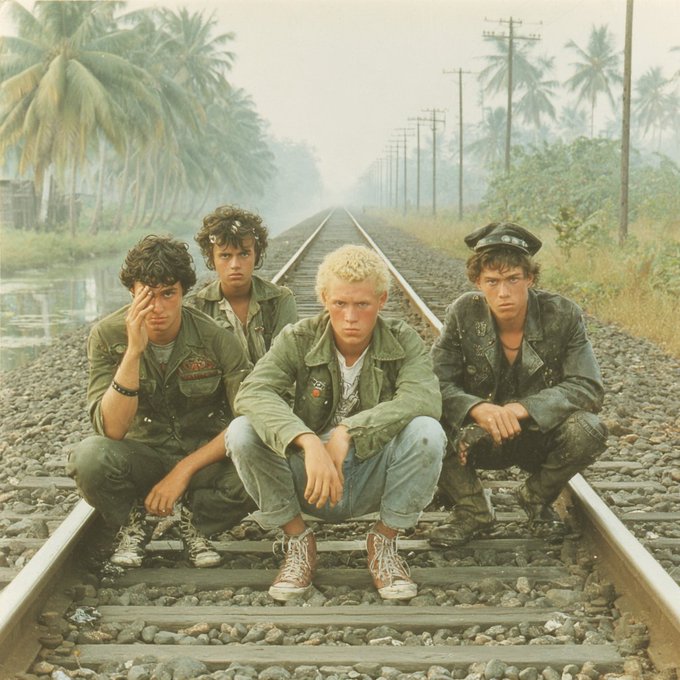 Four young men in military-style jackets and jeans squat on railroad tracks lined with palm trees and power lines in a tropical setting.