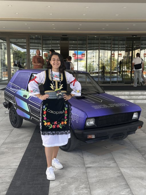A young woman in traditional embroidered attire standing next to a purple Yugo car at what appears to be a car show.