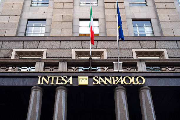 The exterior facade of an Intesa Sanpaolo bank building, featuring stone columns, the bank's gold logo, and the Italian and European Union flags flying above.