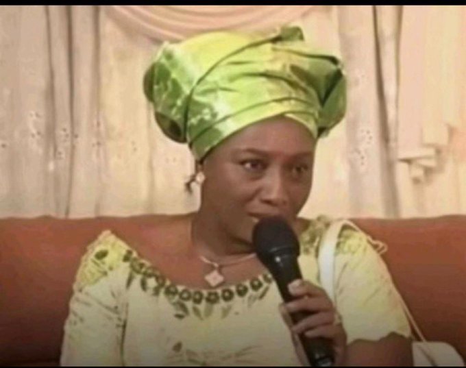 The image features an older woman wearing a green headscarf, sitting down with her face towards the camera while holding a microphone. The background of the photo shows a couch or chair where she is seated. There are also two handbags visible in the scene - one near the woman and another further away from her.