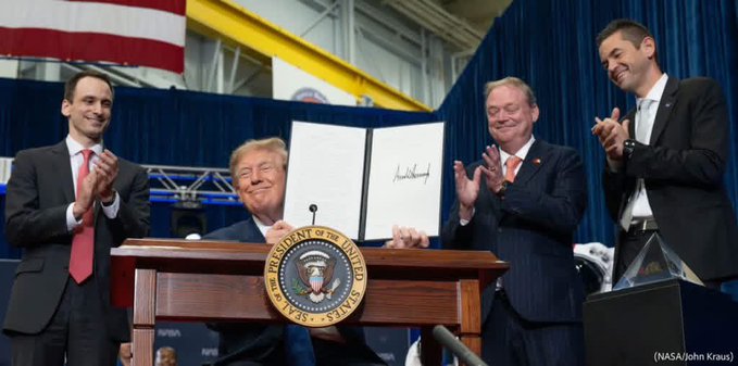 Donald Trump stands at a presidential podium holding up a signed document, flanked by three smiling men in suits who are applauding, with an American flag and a NASA logo visible in the background.