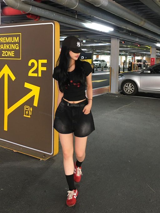 A photo of a young woman wearing a New York Yankees cap, black t-shirt, and black shorts, standing in a parking garage next to a 'Premium Parking Zone 2F' sign.