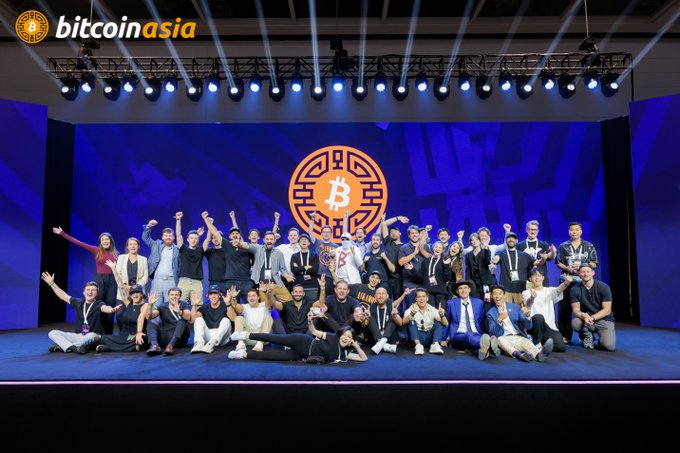 A group photo of people posing on stage at the 'bitcoin asia' event, with a large Bitcoin logo on the screen behind them.