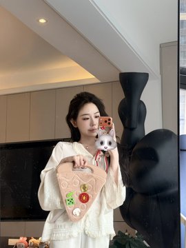 The image features a woman holding up a white purse with a cat design, which has been photoshopped into her photo for fun. The purse appears to be made of fabric or paper mache, giving it a unique appearance. The woman is standing in front of a TV screen and there are several other objects around her, including a potted plant on the left side of the image and another person partially visible behind her.