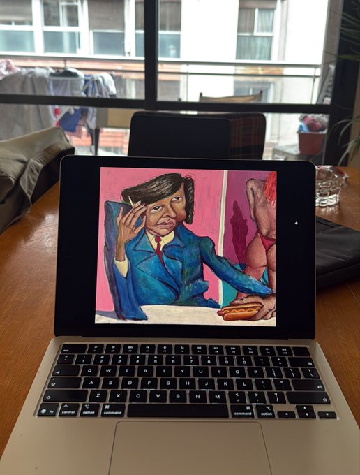 A laptop on a wooden table displays a digital painting of a man in a blue suit holding a pen and a hot dog, with a nude figure visible in the pink background.