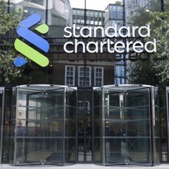 The photo shows the entrance of a Standard Chartered building, featuring the bank's logo and name prominently displayed above glass revolving doors, alongside security booths. This image relates to the post as it depicts the Standard Chartered branch where the bank has launched direct Bitcoin and Ethereum trading services for institutional clients in the UAE.