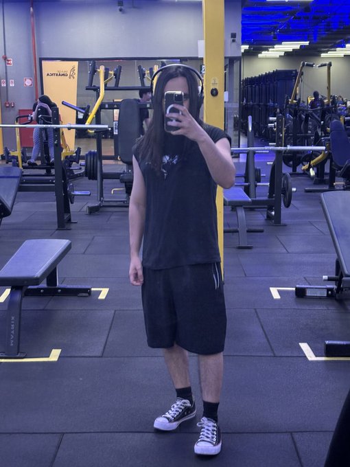 A man in a gym takes a mirror selfie wearing headphones, a dark oversized t-shirt, and black shorts, surrounded by weightlifting equipment. The caption "ANYTIME FITNESS" is visible on a pillar behind him. The image reinforces the post's message about consistency in both fitness and crypto—standing in a gym, headphones on, putting in the daily work while waiting for results.