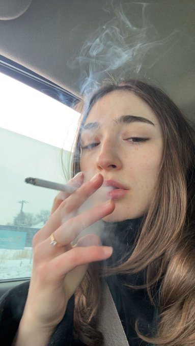 A young woman sits in a car with her eyes half-closed, exhaling a cloud of smoke from a hand-rolled cigarette while wearing a gold ring. The "Gm" caption fits her laid-back, morning-greeting mood with the dreamy, smoke-filled aesthetic.