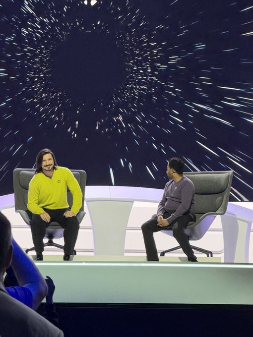 Two men sit on a stage in chairs facing each other for what appears to be a talk show or interview, with a large screen behind them displaying a starfield/warp speed effect. One wears a bright yellow sweater, and the setting suggests a tech or media event where Vlad Tenev (noted in the post) is likely being interviewed.
