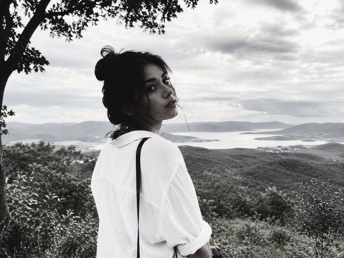 A black-and-white photo shows a woman with her hair in a bun, wearing a white shirt, looking back over her shoulder against a mountainous landscape with a lake in the distance.