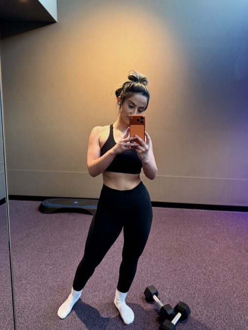 A mirror selfie in a gym showing a woman in black leggings and a sports bra, with her hair in a bun, taking a photo with an orange phone; a dumbbell is visible on the floor.