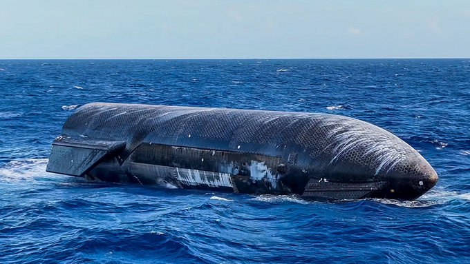 An aerial view of a dark-colored submarine floating partially above the surface of a deep blue ocean.