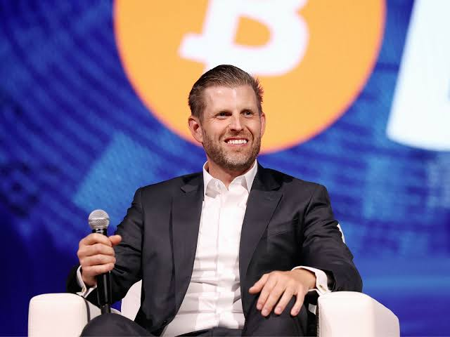 A man with a beard wearing a dark suit jacket and white shirt, smiling while holding a microphone at what appears to be a panel or conference, with a Bitcoin logo visible in the blurred background.