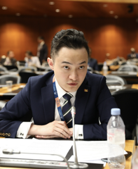 A young man in a dark suit and tie sits at a conference table with a microphone and water bottle, appearing focused in what looks like a corporate meeting or press event. While no text or logos are visible in the image itself, the setting suggests a business or financial announcement context, possibly related to Tron Inc.'s acquisition of additional $TRX tokens as noted in the accompanying post.
