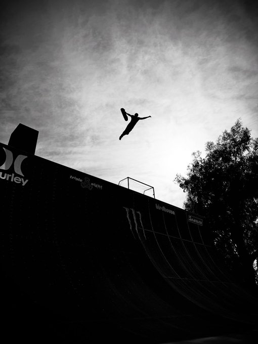 A black and white silhouette photograph captures a skateboarder mid-air performing a trick above a tall vert ramp against a dramatic cloudy sky.