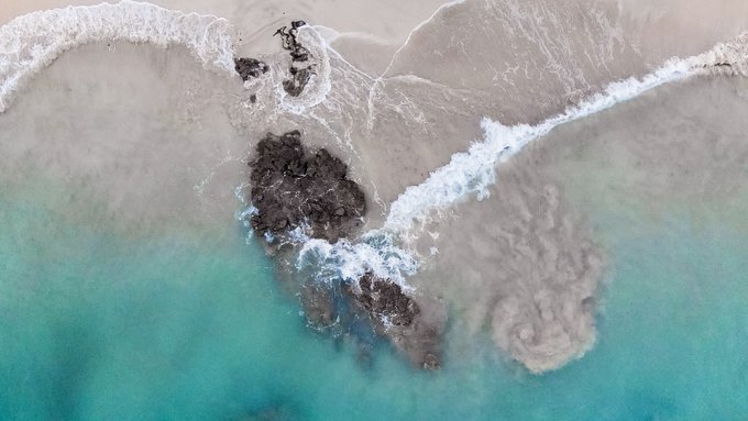 An aerial drone shot captures turquoise ocean waves crashing against dark, jagged rocks jutting out of a sandy shore, with frothy white water swirling around the formations. The image's abstract, otherworldly coastal composition evokes the contemplative uncertainty expressed in the caption about whether to mint this as a unique 1/1 piece or an open edition on Objkt.