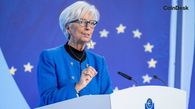 The image features a woman standing at a podium with her hand raised, possibly delivering a speech or presentation. She is wearing glasses and a blue jacket, which stands out against the backdrop of a large blue flag. The flag has stars on it, adding visual interest to the scene. The setting appears to be indoors, as indicated by the presence of a microphone in front of the woman.