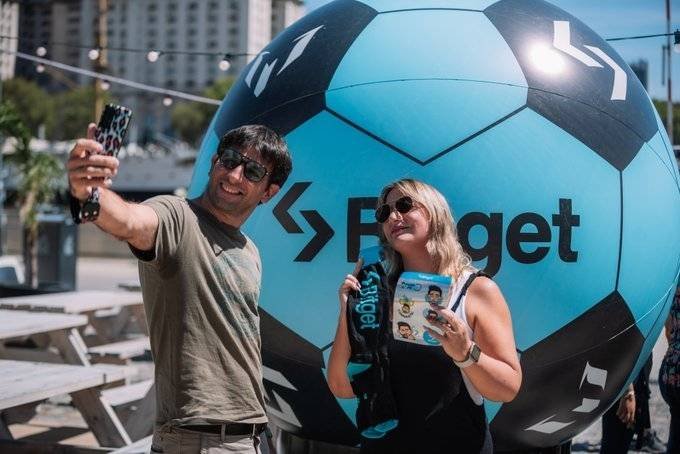 Two people posing for a selfie in front of a large teal and black inflatable soccer ball branded with the 'Fliget' logo at an outdoor promotional event.
