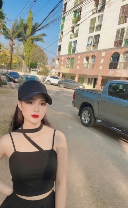 The image features a young woman wearing a black top, standing outside of an apartment building with a truck parked nearby. She has her arms crossed over her chest and is posing for the photo. The surrounding area includes several cars parked along the street, as well as palm trees in the background.