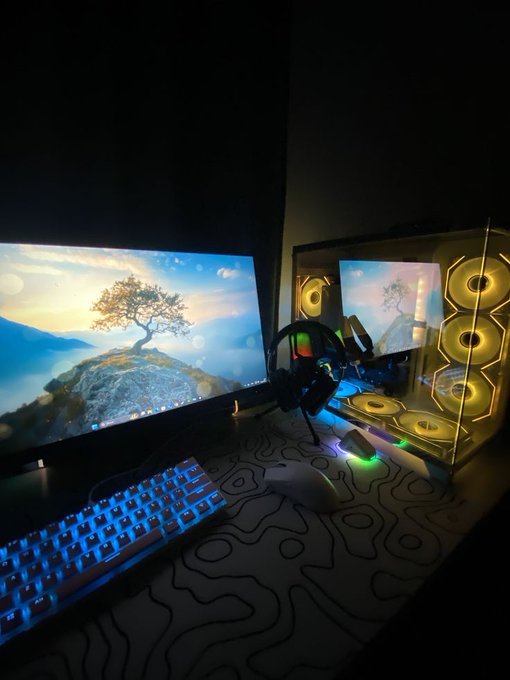 A dimly lit gaming computer setup featuring a dual-monitor setup with a fantasy tree wallpaper, an RGB keyboard on a topographic desk mat, and a PC case with yellow illuminated fans.