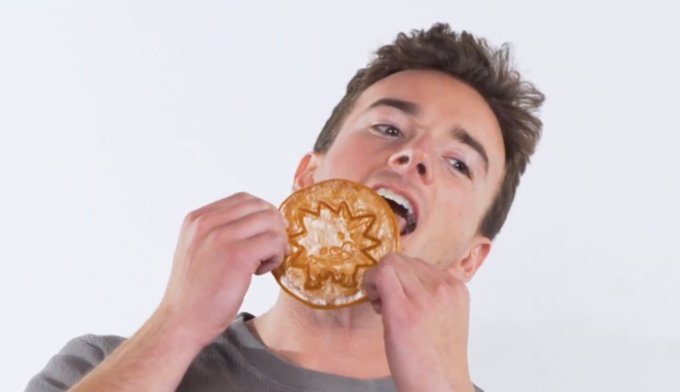 A young man with dark hair enthusiastically bites into a waffle-shaped cookie or pastry, holding it with both hands in front of his face against a plain white background.