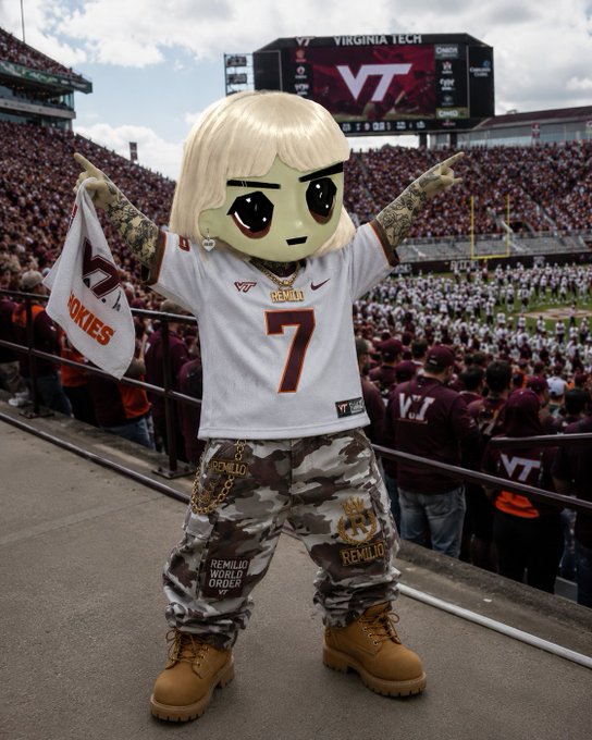 An anime-style mascot character with blonde hair wearing a Virginia Tech football jersey (#7) and camouflage Remilio pants, posing with arms outstretched in front of a crowded stadium scoreboard displaying the VT logo.