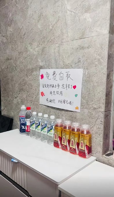 A free beverage station with a handwritten Chinese sign reading '免费自取' (Free to take) thanking travelers, offering bottles of water and red tea drinks on a white counter.