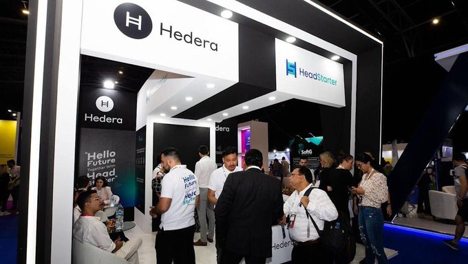 A bustling exhibition booth displays prominent signage for "Hedera" and "HeadStarter" at what appears to be a blockchain or crypto conference, with attendees gathered to network and discuss projects. The branding directly connects to the post's mention of $HBAR and HeadStarter's HST token, showcasing a real-world event where the Hedera ecosystem and HeadStarter platform are engaging with potential users and investors.