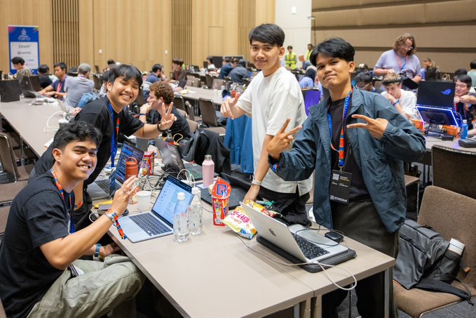 Several young men pose cheerfully at a table with laptops, water bottles, and snacks, flashing peace signs in what appears to be a gaming or tech event.
