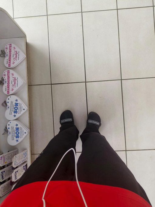 First-person view looking down at feet wearing black Columbia shoes, black pants, and a red top, standing on white tiled flooring next to a display of branded guitar picks (Granddaughter and NOS).