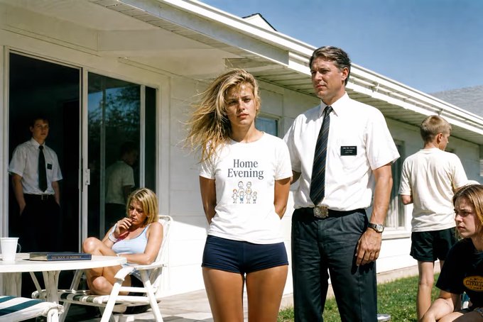 A scene depicting a young woman in a 'Home Evening' t-shirt and dark shorts standing next to a man in a white shirt and tie with a nametag reading 'Elder Price', with other similarly dressed people around them.