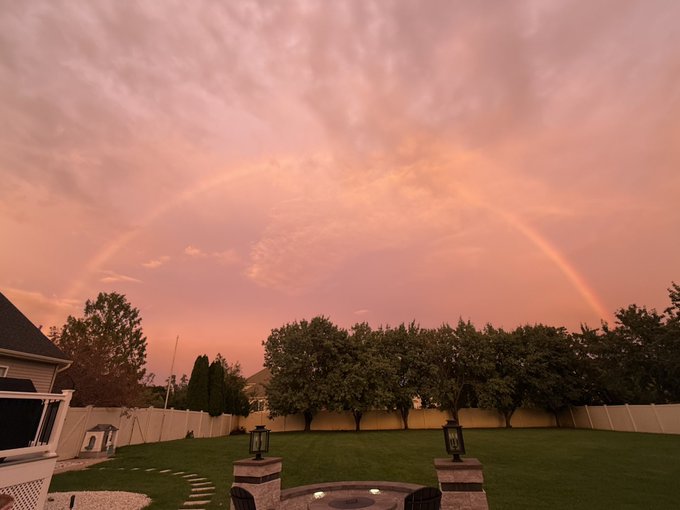 A rainbow arcs across a pink-hued sunset sky over a backyard with green trees, a white fence, and a stone patio.