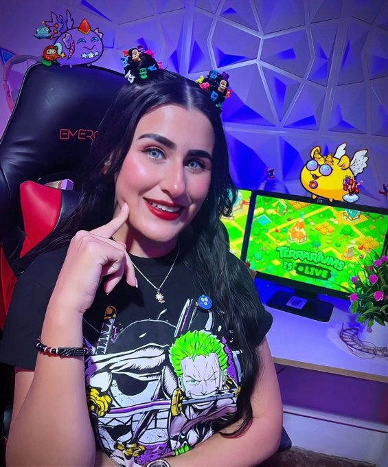 The image features a woman wearing a black t-shirt with a cartoon character design. She has her hand on her face and is smiling at the camera. The background of the photo shows a TV screen displaying a video game, indicating that she might be playing or watching a gaming session. There are also two potted plants in the scene, one near the woman's head and another further away from her.