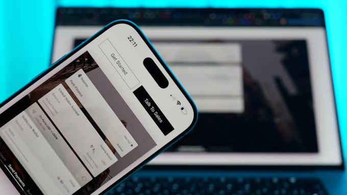 A smartphone displays a 'Get Started' onboarding screen in front of a laptop, showing the time 22:11 with options including 'Help & Support' and 'Talk To Sales'.