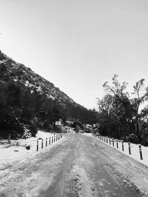 A black and white photograph of a snow-covered road lined with short posts, leading toward snow-dusted hills and trees.