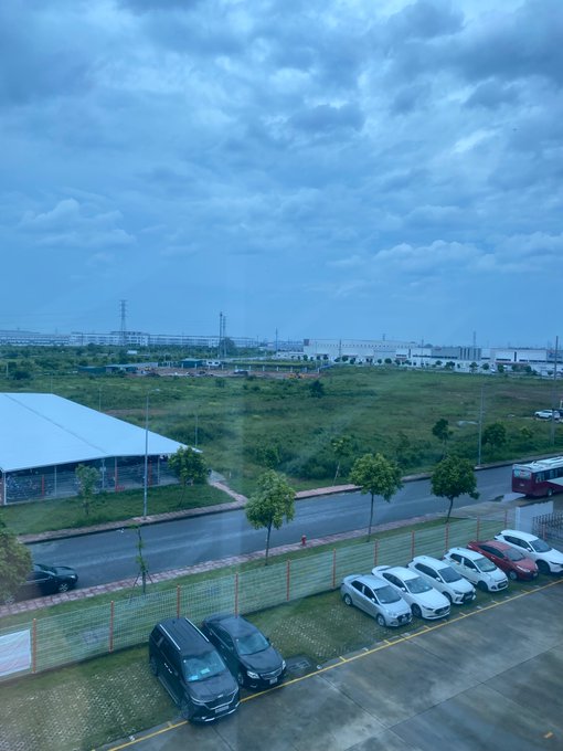 An overcast landscape view from an elevated window showing a parking lot with several cars, a fenced road, grassy fields, and industrial buildings in the distance.