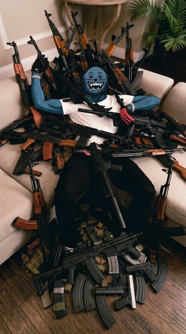 A person wearing a blue smiling face mask and white t-shirt sits on a couch surrounded by an enormous pile of various firearms, magazines, and ammunition.