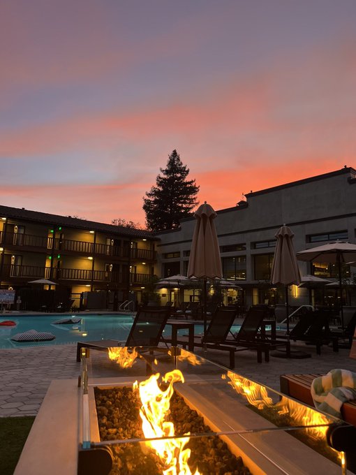 A serene poolside scene at sunset shows a fire pit in the foreground, lounge chairs, closed umbrellas, and a multi-story building under a pink and orange sky.