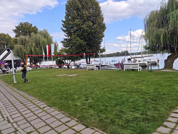 A person plays volleyball on a grassy area near a waterfront with sailboats, willow trees, and outdoor seating in the background.