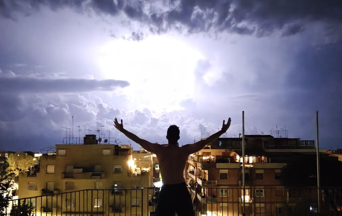 A silhouetted shirtless man stands on a balcony with arms raised triumphantly toward a dramatic, brightly illuminated cloudy sky over an urban cityscape at night.