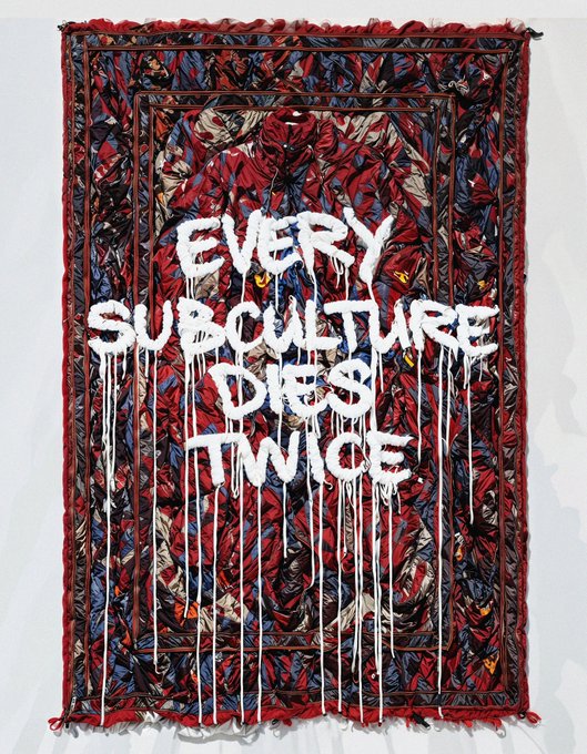 A frayed, ornately patterned red and blue Persian-style rug features the dripping white graffiti text "EVERY SUBCULTURE DIES TWICE," playing on the phrase "carpe diem" by suggesting subcultural rebellion itself becomes commodified and co-opted, much like a carpet transformed from functional object into decorative display.