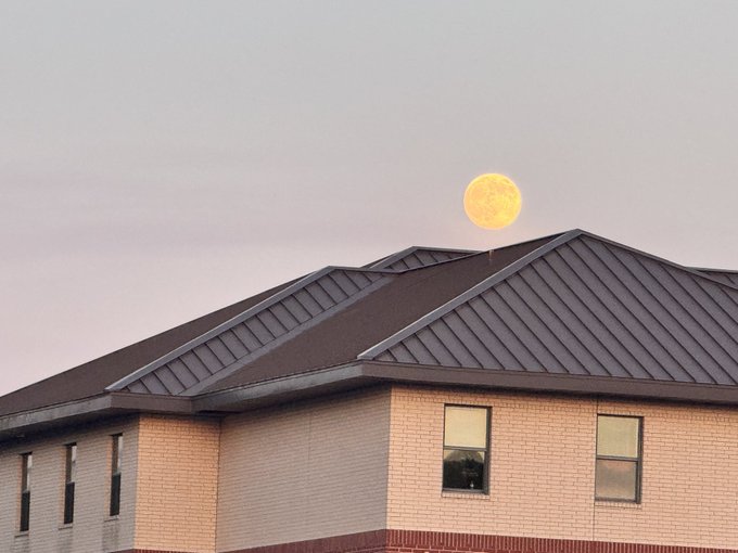 A large yellow full moon setting behind the dark gray shingled roof of a brick building at dusk.
