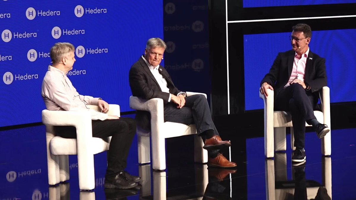 The image shows three men sitting in chairs, likely at a conference or event. They are positioned behind a blue screen with white text, which appears to be part of a presentation or discussion. The background features a large screen displaying the words "Hedera" and "Hedera", suggesting that this gathering is related to the company's name or vision. The men seem engaged in conversation, possibly discussing the ideas presented on the blue screen.