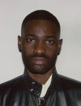 A close-up portrait of a man with a beard wearing a black leather jacket over a patterned shirt against a light background.