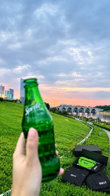 A hand holding a green Sprite bottle in the foreground against a scenic sunset view of a park with a bridge over water, city skyscrapers in the distance, and 'OKX' branded merchandise displayed on the grass.