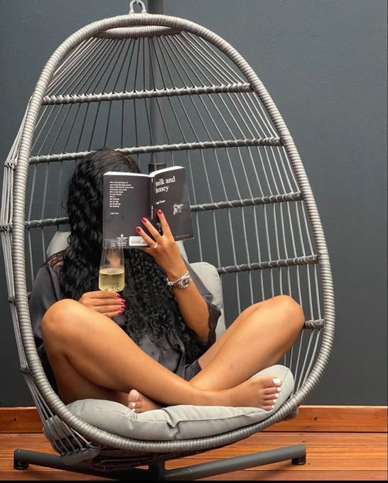 A woman sitting cross-legged in a gray hanging egg chair, reading a book titled 'milk and honey' while holding a glass of white wine.