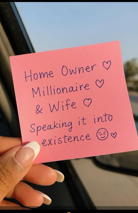 A hand holds a pink sticky note with handwritten text reading 'Home Owner ♡ Millionaire ♡ & Wife ♡ speaking it into existence :) ♡'.