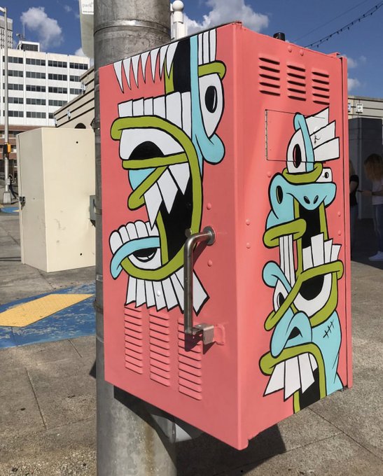 A pink utility box on a city street decorated with vibrant street art featuring multiple stylized blue and green eyes with sharp teeth, set against an urban backdrop with buildings and a blue sky.